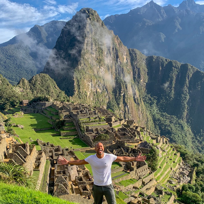 Jermaine with his arms outstreched at the ancient city of Machu Picchu with a scenic mountain backdrop